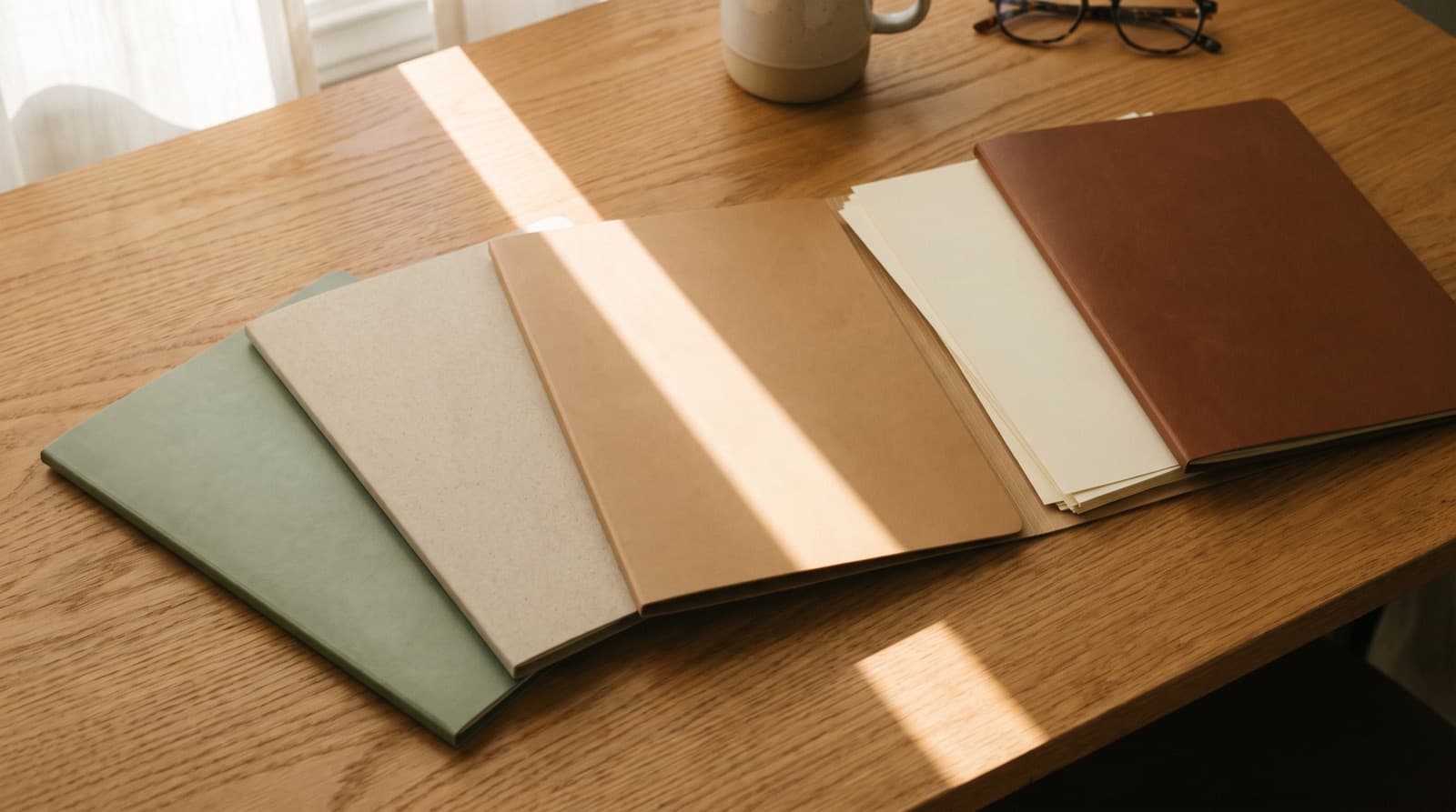 Four slim document folders in muted sage, oatmeal, tan and cognac fanned across a sunlit oak desk.