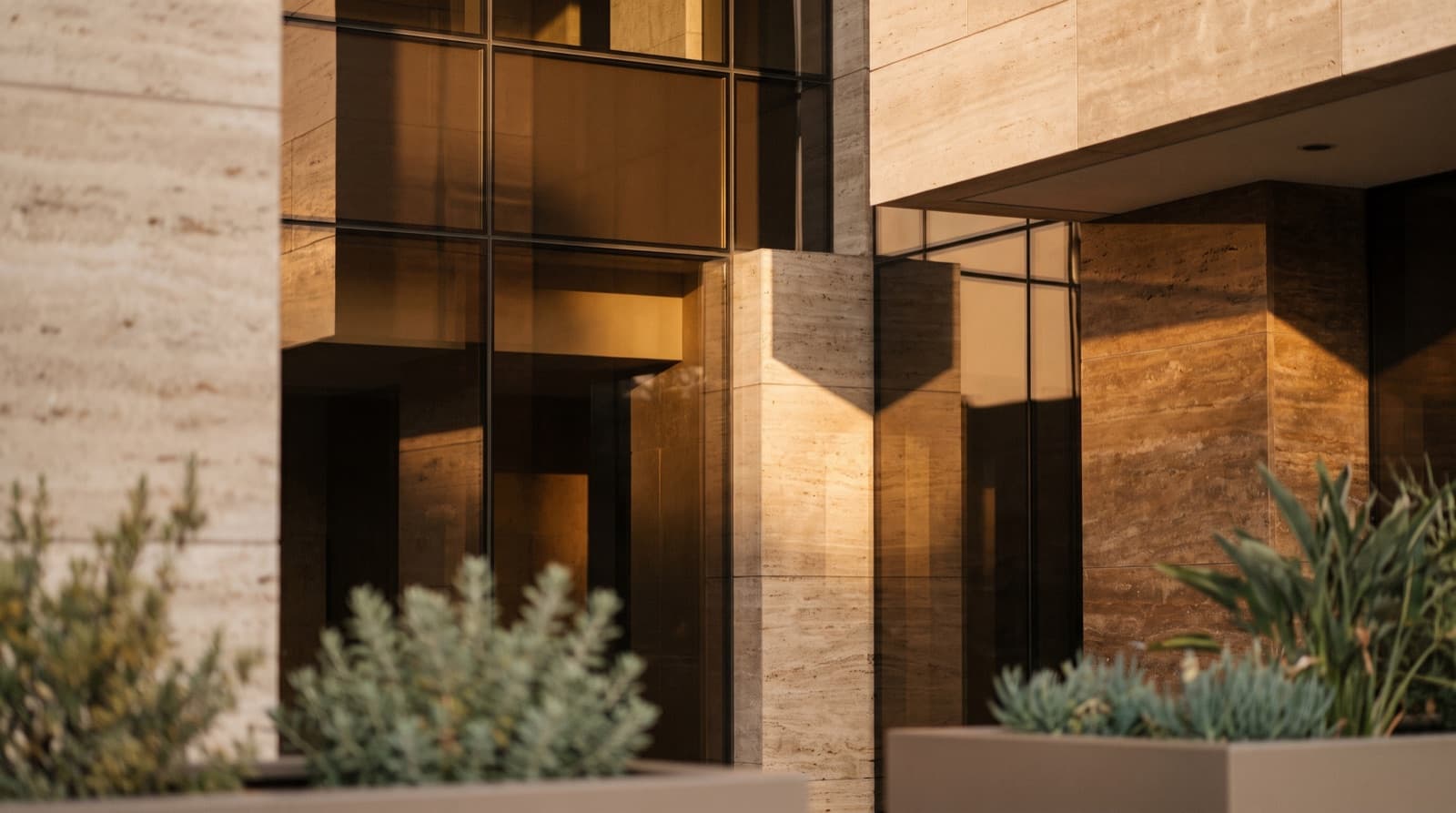 Intersecting planes of bronze glass and warm travertine on a contemporary facade, lit low and raking.